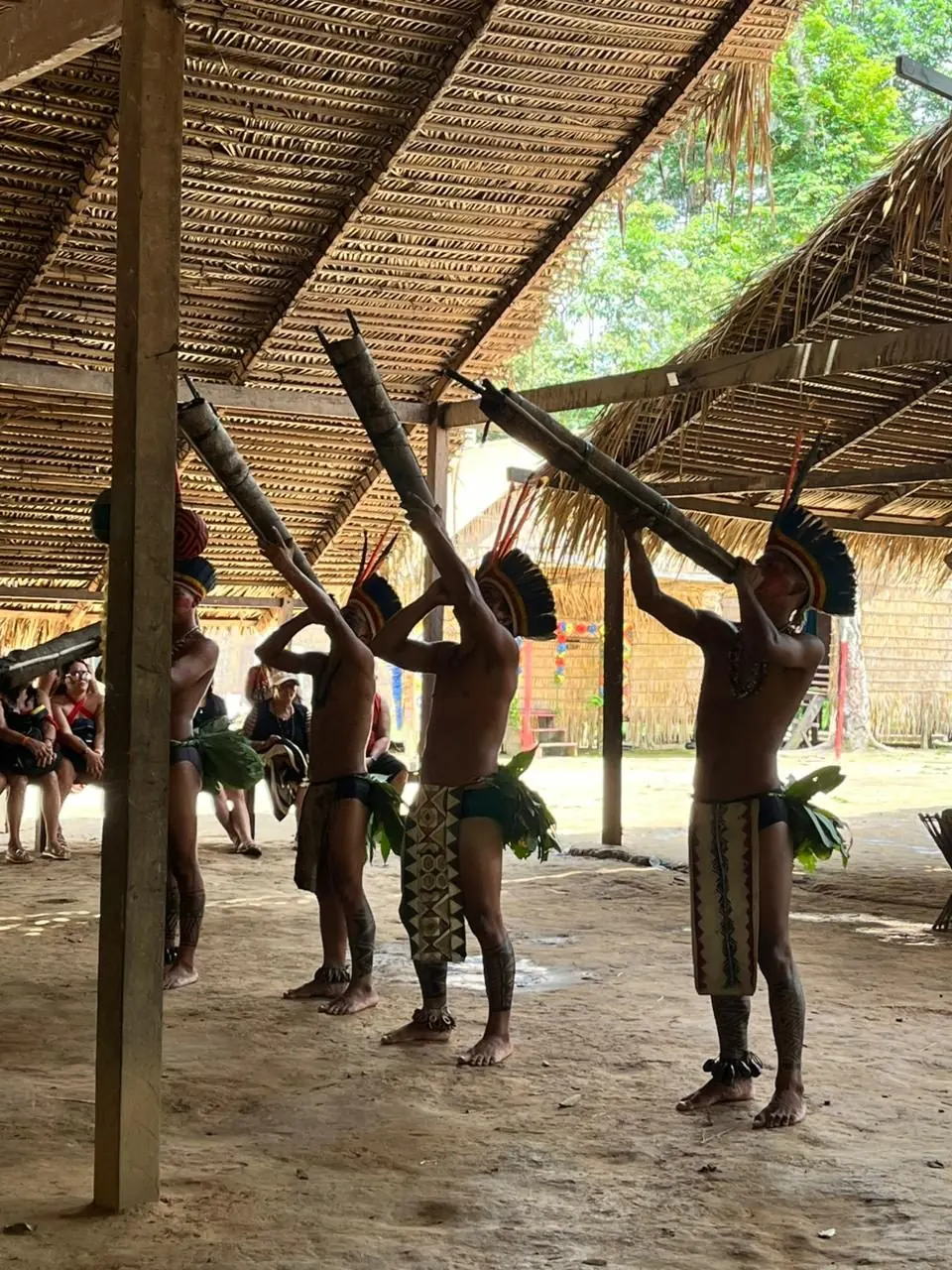 Indigenous dancers in feather headdresses playing traditional wind instruments inside the village maloca